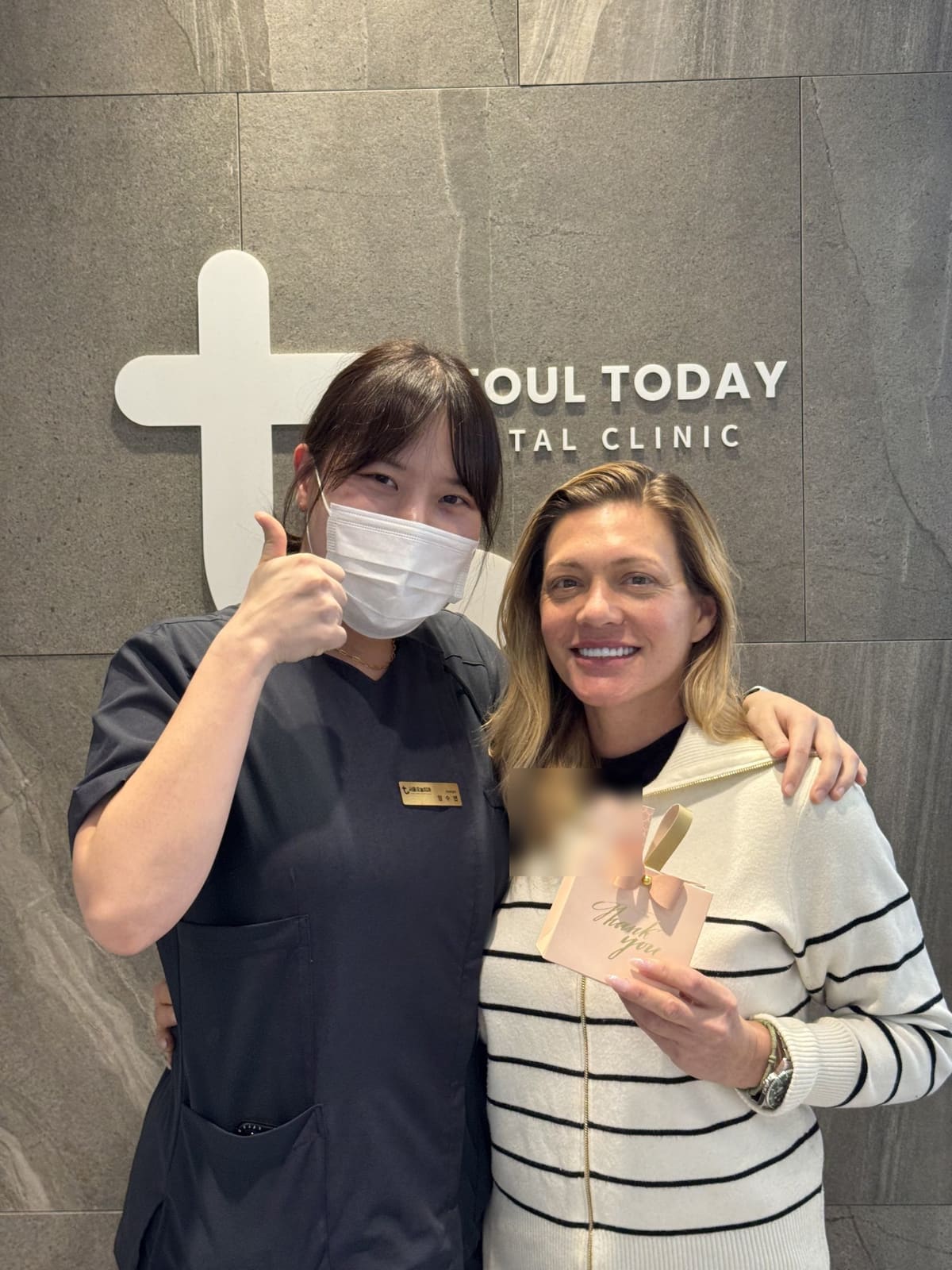 A smiling international patient holding a thank-you gift bag with a staff member in front of the Seoul Today Dental Clinic logo wall